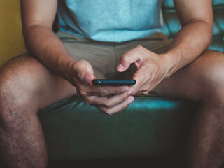 Hand man holding smartphone. A young person, man in casual grey shirt and shorts sitting and interacting with technology at home. Relaxation male using mobile phone.