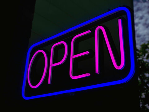 OPEN neon sign on glass at the entrance to bar or cafe, dark background, illuminated pink text in a blue square sign saying "OPEN", for business sign.