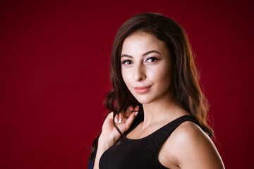 portrait of a young attractive adult brunette woman with long hair in a black short top on a dark red background. The model looks at the camera and smiles. Close-up.