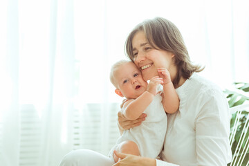 Young mother hugging her little daughter, they sitting near window and smiling. Concept of happy family, motherhood, womens or mother day