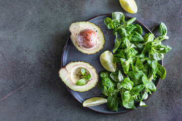 Avocado, green leaves salad and lime slices on green concrete background. Healthy fats. Top view. Copy space.