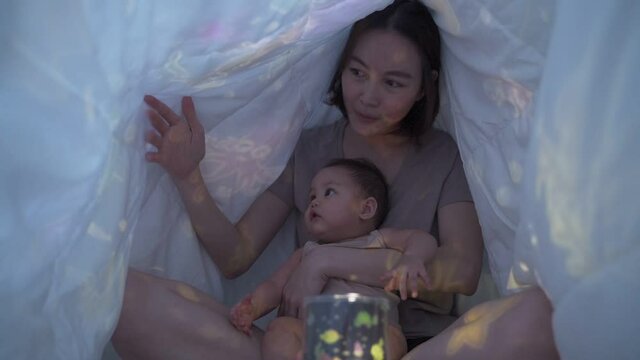 asian woman mother parent playing light projector toy with baby infant under blanket in bedroom at home. pointing picture shadow teaching baby a lesson before bed time.
