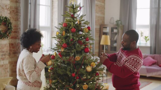 Positive Afro-American family couple decorating Christmas tree together and chatting while preparing home for celebration