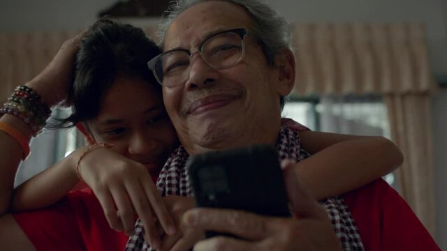 Asian Senior Gray Hair Grandfather Watching TV From Mobile Smartphone In Living Room While His Adorable Granddaughter Approach And Hug Him From Behind Happily. Bonding Relationship In Family.