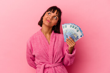 Young mixed race woman wearing a bathrobe holding bills isolated on pink background dreaming of achieving goals and purposes