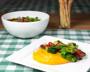 Beef medallions with pea puree served on a square white plate with a bowl of salad on table. Over green plaid tablecloth