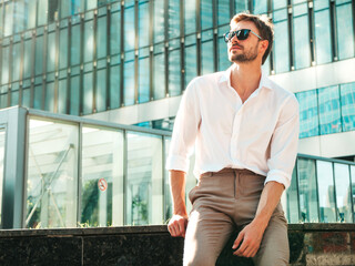 Fototapeta premium Portrait of handsome confident stylish hipster lambersexual model.Modern man dressed in white shirt. Fashion male posing in the street background near skyscrapers in sunglasses