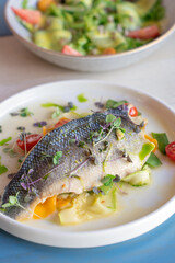 Roasted cooked half of fish decorated by chef with cherry tomatoes, cucumber slices, microgreen and green sauce served on white round plate on wooden table at restaurant. Mediterranean food, vertical