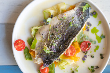 Top view of fried cook half of fish decorated by chef with cherry tomatoes, cucumber slices, microgreen and green sauce served on white round plate on wooden table at restaurant. Horizontal image