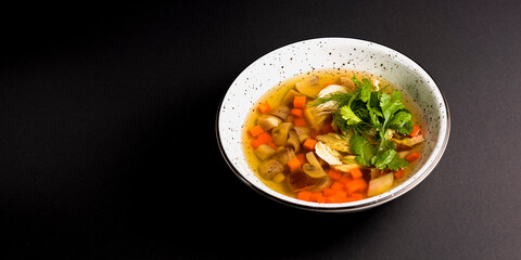 Chicken soup with vegetables amd mushrooms, fresh parsley and dill on top. Served in a white bowl over black background