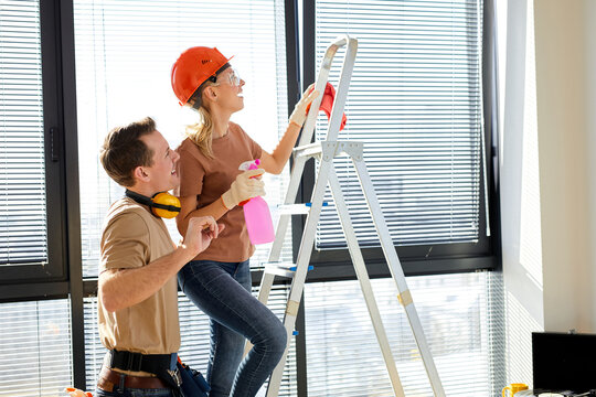 Helpful man and woman cleaning house after renovation, man help wife, use stepladder for better cleaning of ceiling, holding spray bottle in hands, wearing hardhat. side view