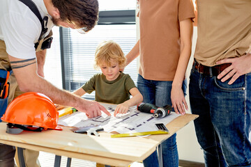 Friendly builder in uniform explain repair plan of new house to customers clients, showing plan on...