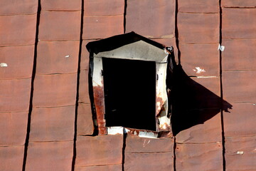 Twisted damaged broken old roof window frame without glass on top of abandoned suburban family house roof covered with rusted cracked metal roof tiles on warm sunny spring day