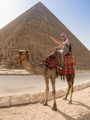 A tourist on a camel poses against the backdrop of the pyramids in Giza, Cairo, Egypt. Kufiya is wrapped around the man's head. The camel stands in full growth.