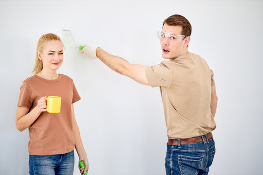 Funny caucasian married couple paint the wall, woman have rest, while husband is painting, looking at side, in safety glasses. side view portrait
