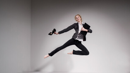 Graceful barefoot ballerina in a business suit jumping with shoes in her hands on a white background.