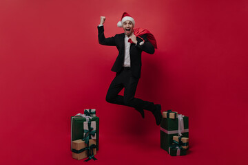 Happy young boy in light shirt, black suit and bright Santa hat jumping with bag near stacks of gift boxes, shouting against red plain background 