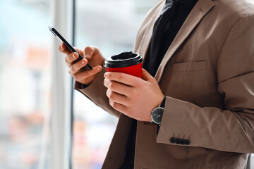 Young man with cup of coffee using mobile phone in office