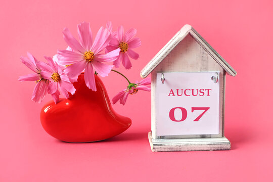 Calendar for: August 7 : the name of the month of August in English with the numbers 0 and: 7 on a toy house, a bouquet of pink flowers in a heart-shaped vase, pink background