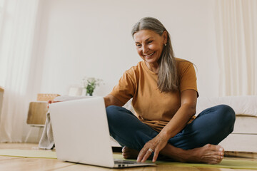 Indoor image of beautiful energetic female on retirement sitting barefoot on floor using laptop turning on yoga video tutorial. Elderly European woman surfing internet on portable computer