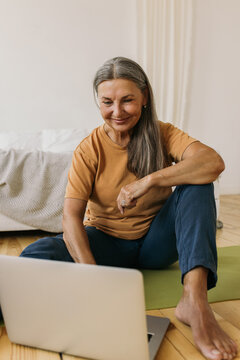 Technology, Gadgets And Aging Concept. Barefoot European Female Pensioner Using Portable Computer At Home, Siting On Carpet, Surfing Internet Or Watching Online Yoga Video Tutorial For Aged People