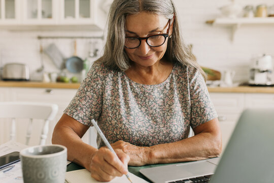 Mature concentrated woman writing down new ideas in notebook sitting on kitchen. Cozy atmosphere with cup of coffee. Inspiration, creativity, writer, freelance job concept