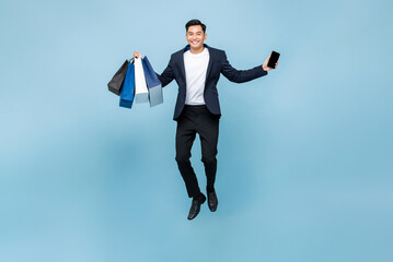 Excited young handsome Asian man holding mobile phone and shopping bags jumping in light blue...