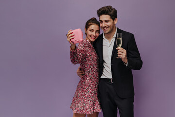 Cute pair of young people standing in party outfits, smiling and making selfie against light purple background. Man holding glass of champagne 