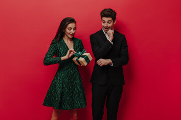 Handsome young man in stylish black suit waiting for his girlfriend in green dress to unpack gift box fascinatingly. Red plain background 