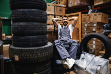 Fototapeta na wymiar Smiling lazy tattooed bearded worker sitting on tires in storage of import and export firm.