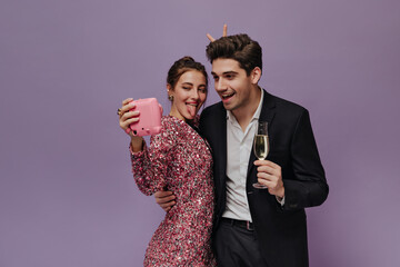 Joyful young girl in pink glitter dress making selfie with boyfriend, wearing white shirt, black suit and holding glass of champagne. Couple posing isolated on violet background 