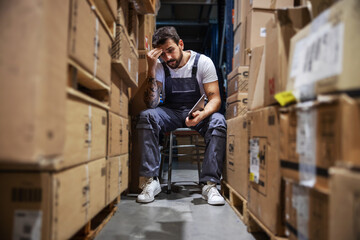 Young worried hardworking tattooed bearded blue collar worker in overalls sitting on the chair in...