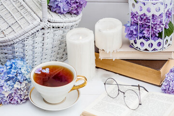 A visual for content. Still life in vintage style. A mug with a drink, an old book, a cage, candles and hydrangea flowers in the garden on a white wooden table. The concept of a tea ceremony.