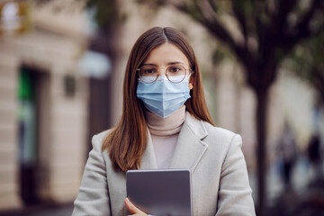 Brunette with face mask standing on the street and holding tablet during corona virus.