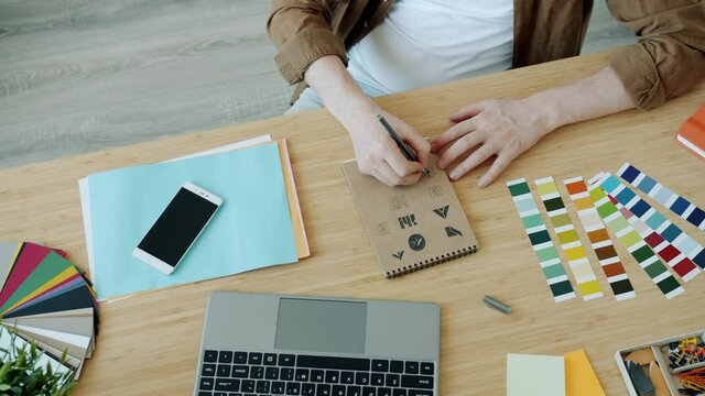Close-up slow motion of male hands drawing logo design on paper and using laptop typing indoors in office. Creative activity and modern technology concept.