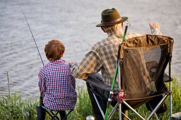 View from back on nice senior man with grandson sitting near river with fishing rods in hands, enjoying beautiful nature, little boy learn to fish with grandfather. at weekends in evening, at sunset