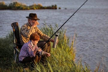 Glad grey-headed man with grandson on fishing, drinking tea from thermos, have rest in nature, leisure time, enjoy and relax. Side view portrait of family in checkered shirt on lake in the evening