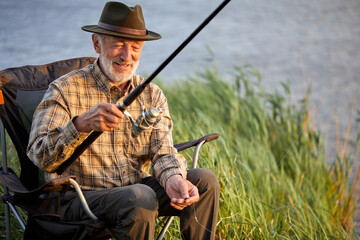 Elderly happy man fishing outside in evening on lake in summer sitting on chair, enjoy spending time in nature, want to catch a fish, alone, side view portrait of male in casual checkered shirt