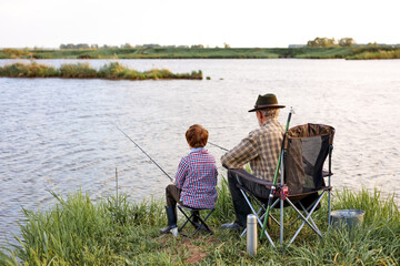 Back view portrait of adult man and teenage boy sitting together on lake fishing with rods in calm waters of blue lake in countryside, both wearing checkered shirts