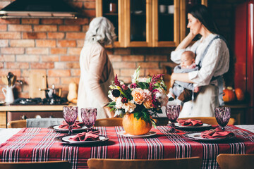 Young woman with long loose hair holds baby in sling bag and helps aged mother serving large table with plates and autumn flower bouquet in kitchen.