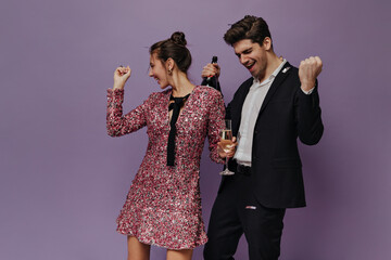 Couple of attractive young people in party outfits dancing and celebrating with champagne. Violet plain background 
