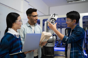 Caucasian male engineer standing hold a robot controller next to Asian female apprentice holding a laptop, demonstrating the work of robotic machine to young Asian male CEO who smile touching a robot. - obrazy, fototapety, plakaty