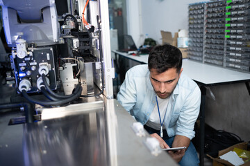 Young Caucasian male electrical engineer in blue shirt squatting at the back of machine checking wire cables and electrical circuit of the machine with digital tablet in hand in an industrial factory