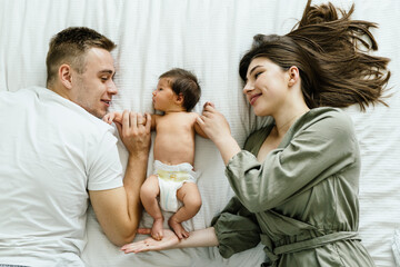 Happy family, husband and wife lying on bedding near adorable newborn baby in the middle of them. Parenting, love, caressing hands from first days of life concept.