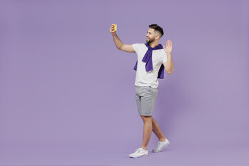 Full size body length cheerful young brunet man 20s wears white t-shirt purple shirt move go do selfie shot on mobile cell phone meet greet wave hand isolated pastel violet background studio portrait