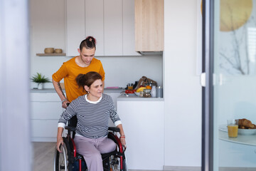 Adult son helping disabled mature mother in wheelchair in kitchen indoors at home.