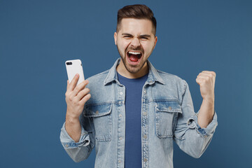 Excited young brunet man 20s wears denim jacket hold in hand use mobile cell phone talk speak on...