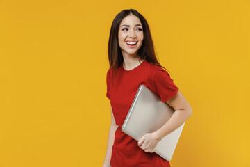 Smiling excited vivid charming fun happy cheerful young brunette woman 20s wears basic red t-shirt hold under hand laptop pc computer looking back behind isolated on yellow background studio portrait.