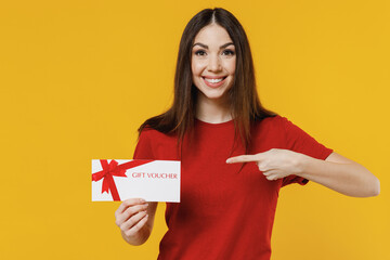 Smiling excited vivid overjoyed fascinating young brunette woman 20s wears basic red t-shirt hold point on gift certificate coupon voucher card for store isolated on yellow background studio portrait - obrazy, fototapety, plakaty
