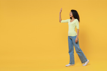 Full size body length side view profile happy african american young woman 20s wears green shirt move go step meet greet waving hand as notices someone isolated on yellow background studio portrait